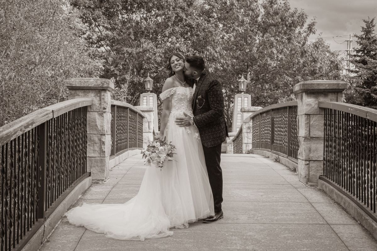 A bride and groom share an intimate kiss on a stone bridge, surrounded by soft, diffused light filtering through trees.