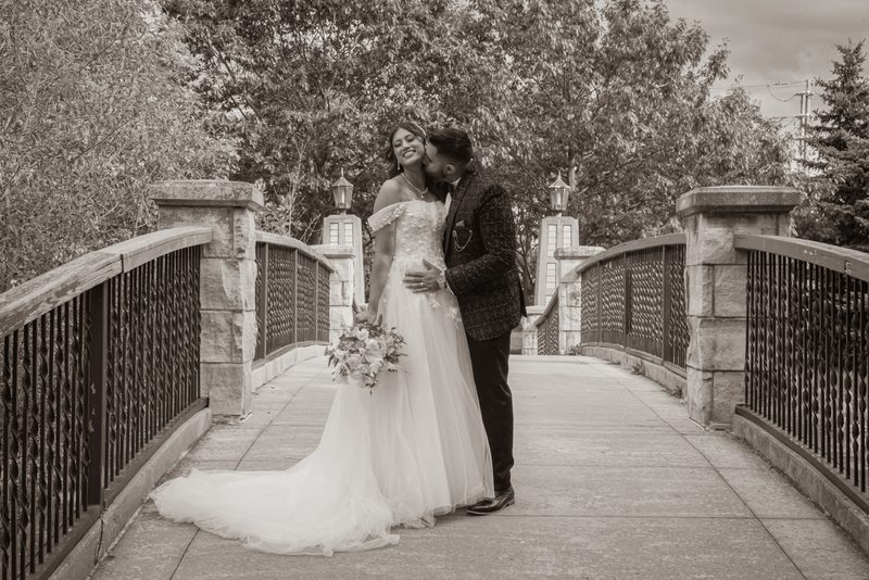 A bride and groom share an intimate kiss on a stone bridge, surrounded by soft, diffused light filtering through trees.