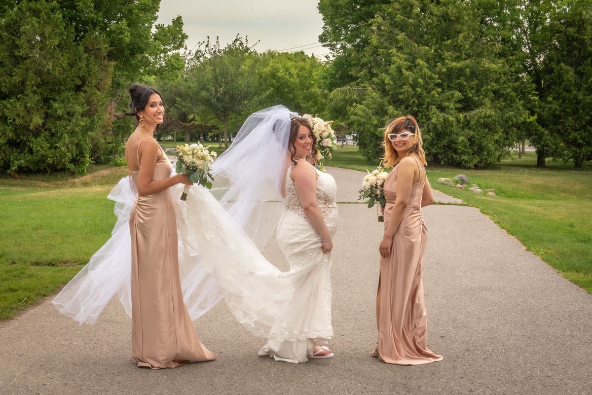 A bride in a lace gown and veil stands with two smiling bridesmaids holding floral bouquets on a grassy, tree-lined path.