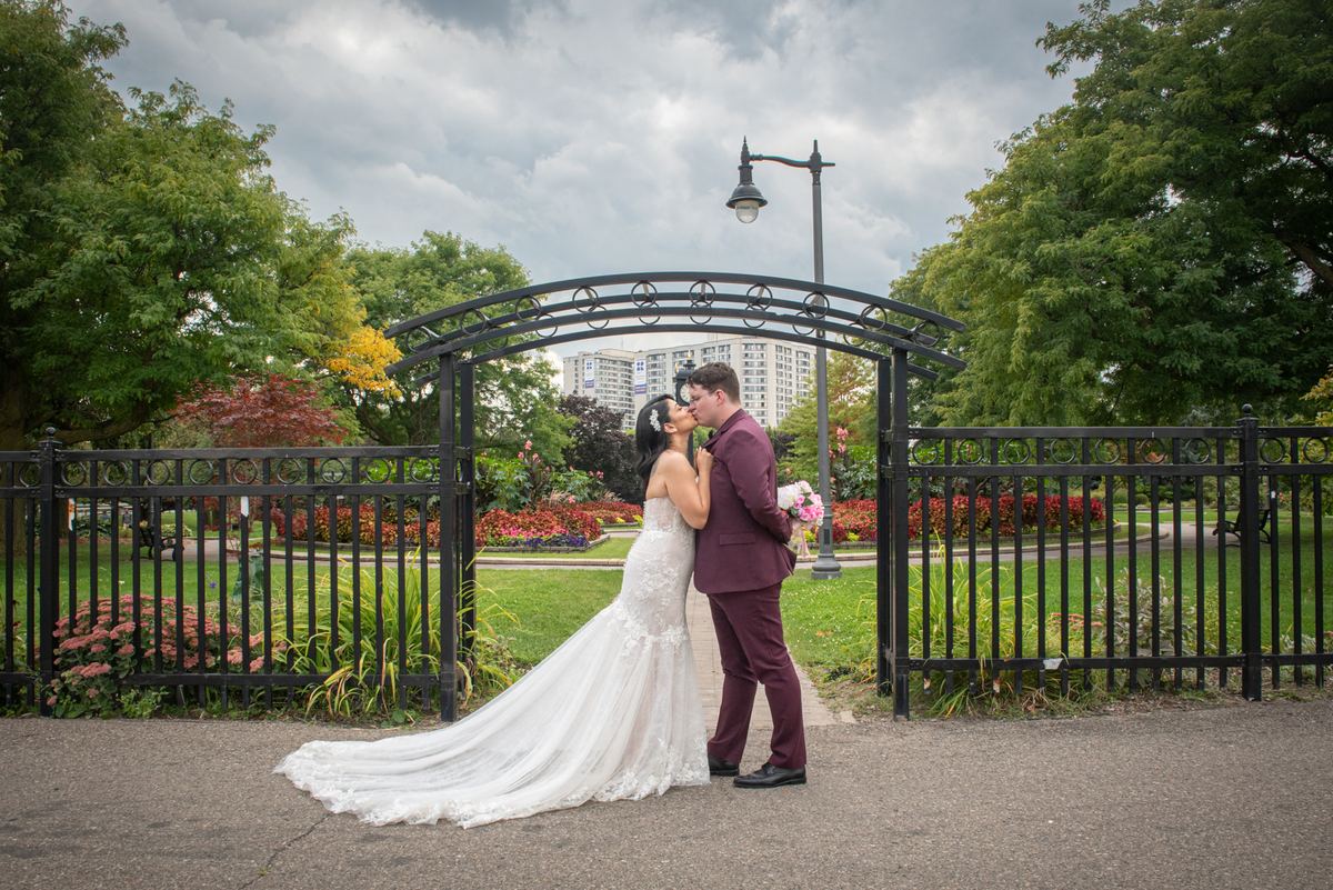 A bride and groom kiss beneath a black iron arch in a lush park, framed by vibrant flowers and trees.