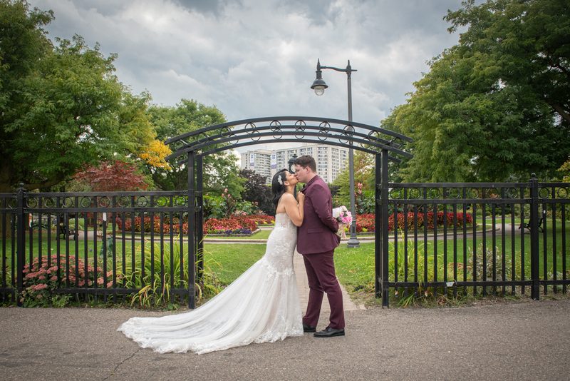 A bride and groom kiss beneath a black iron arch in a lush park, framed by vibrant flowers and trees.