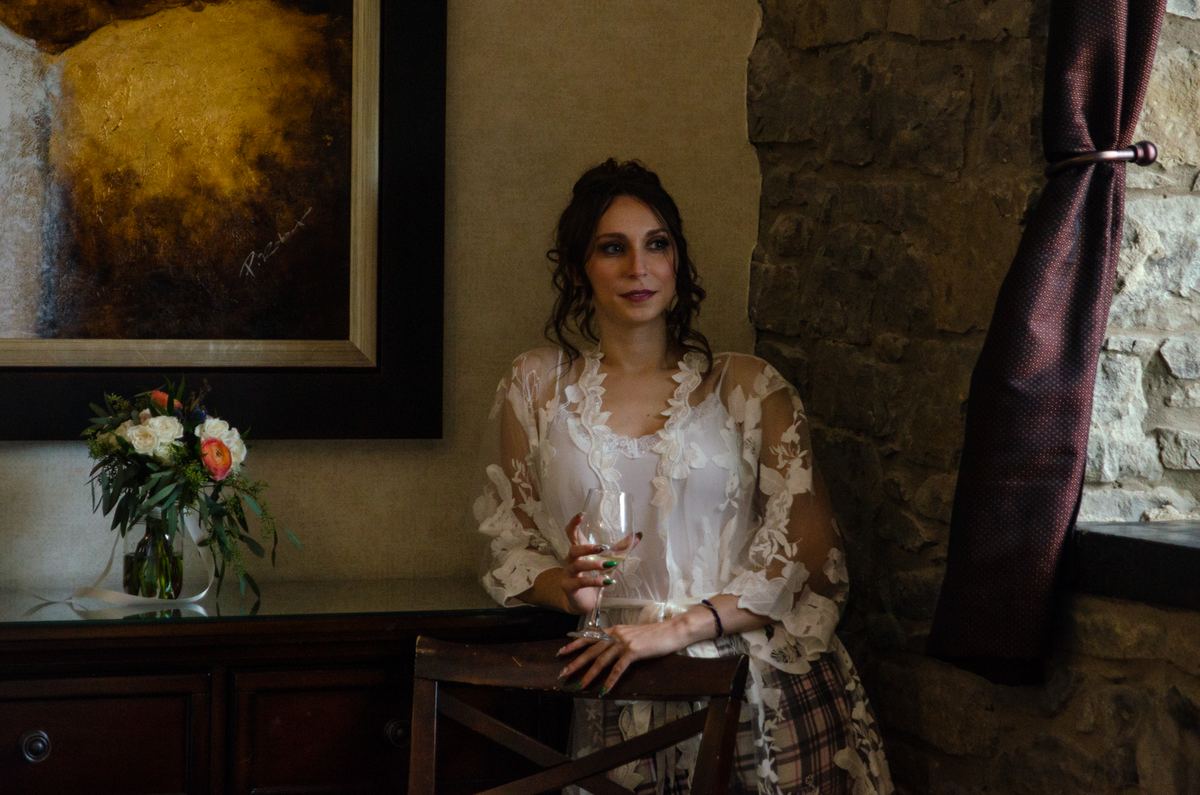 A bride in a lace robe leans against a rustic stone wall, holding a wine glass beside a dark wooden console with flowers.