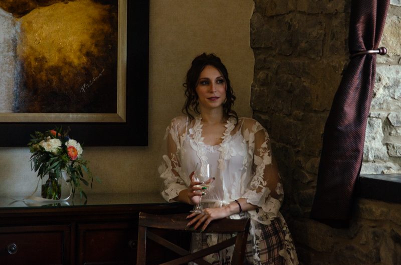 A bride in a lace robe leans against a rustic stone wall, holding a wine glass beside a dark wooden console with flowers.