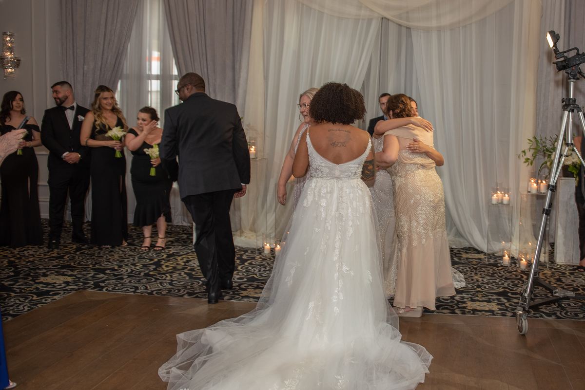 A bride in a white gown with a tattoo on her back is embraced by a woman as they walk toward the altar, guests watching.
