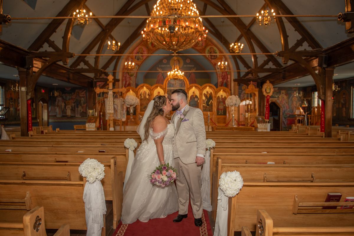 Bride and groom kiss at an Orthodox church altar, surrounded by wooden pews and white flowers, under golden chandeliers.