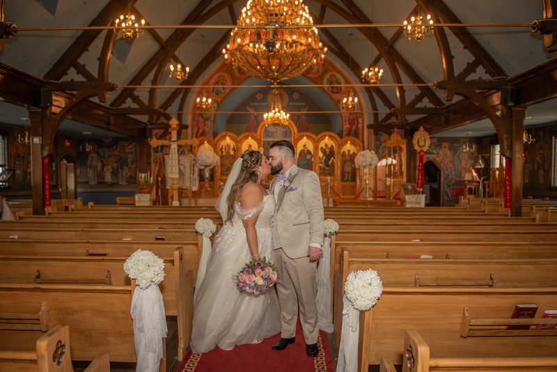 Bride and groom kiss at an Orthodox church altar, surrounded by wooden pews and white flowers, under golden chandeliers.