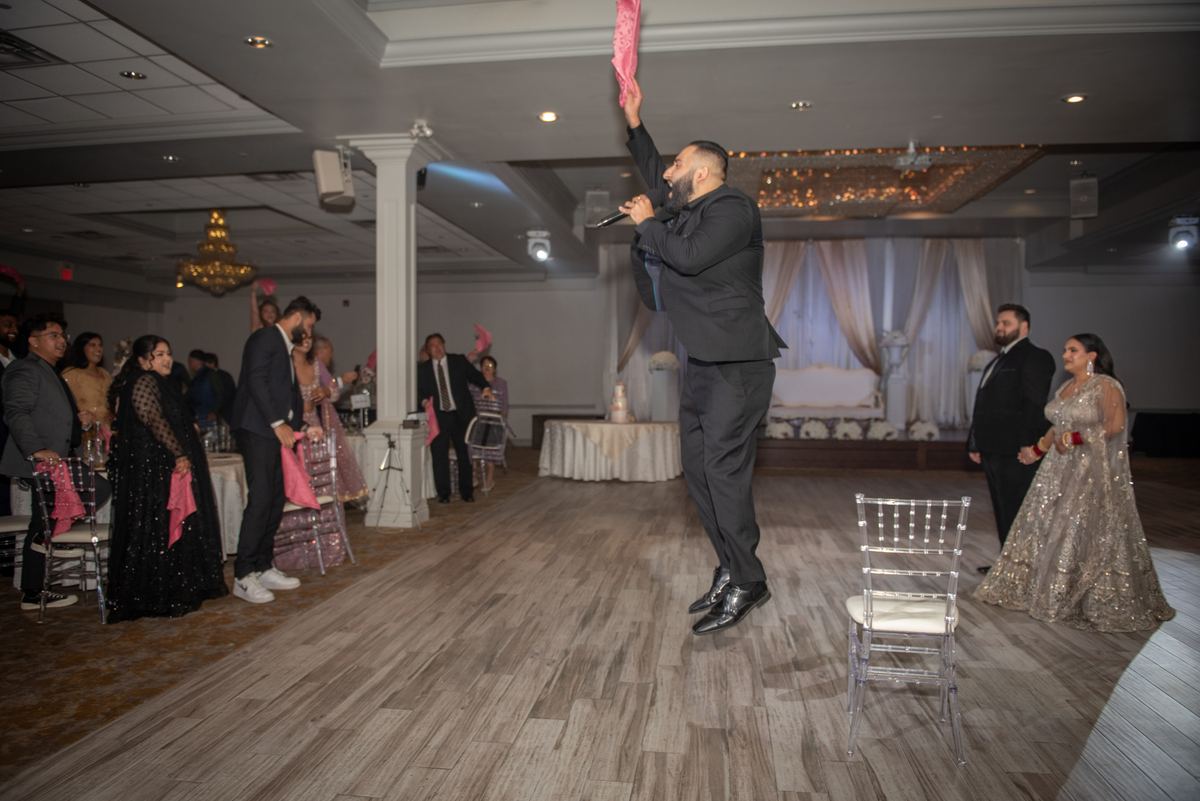 A man in a black suit holds a pink cloth high while singing on stage at a formal celebration, surrounded by guests.