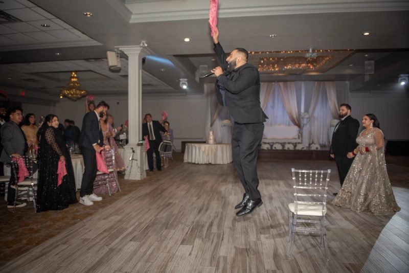 A man in a black suit holds a pink cloth high while singing on stage at a formal celebration, surrounded by guests.