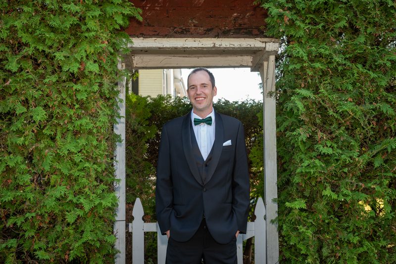 A man in a black tuxedo with a green bow tie stands by a white picket gate and lush hedges under warm afternoon light.