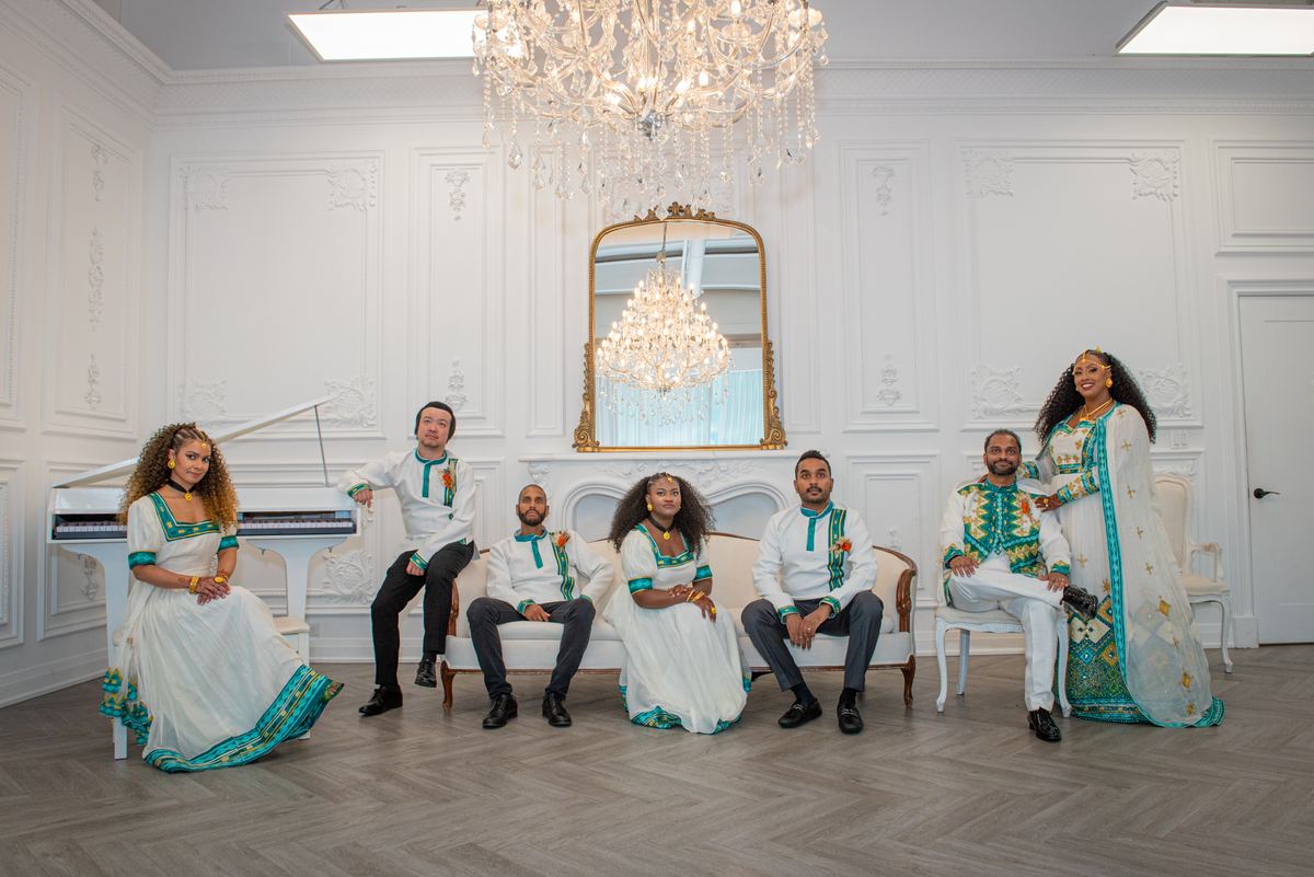 Seven people in elegant traditional attire pose in an opulent room with a crystal chandelier and grand piano.