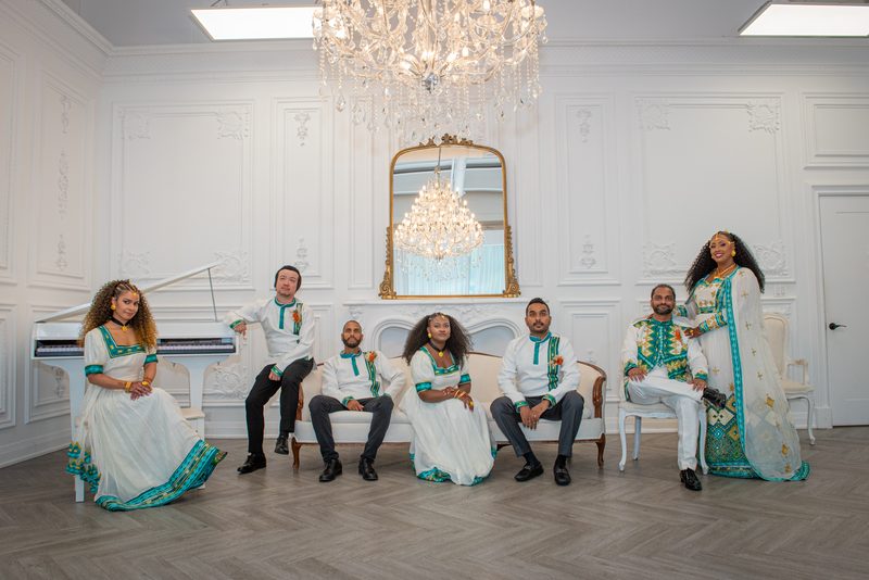Seven people in elegant traditional attire pose in an opulent room with a crystal chandelier and grand piano.