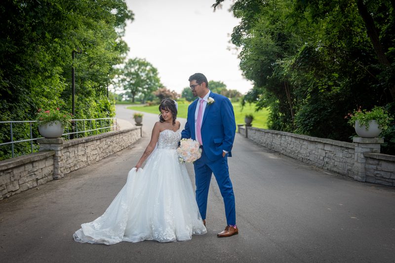 A bride and groom stand on a paved path, surrounded by stone walls and greenery, in an intimate moment.