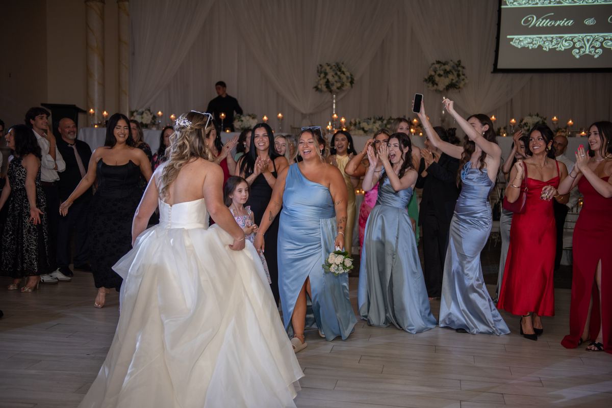 Bride in white gown walks with bridesmaid in blue dress through cheering crowd at candlelit celebration.