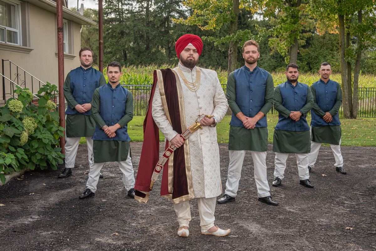 A Sikh groom in white and red stands with his groomsmen in matching attire beside a house under diffused daylight.