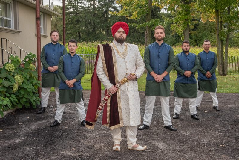 A Sikh groom in white and red stands with his groomsmen in matching attire beside a house under diffused daylight.