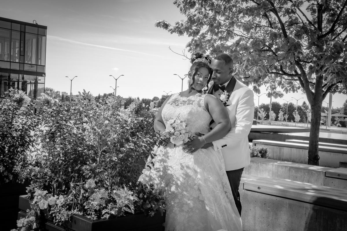 A bride in a lace gown and tiara is embraced from behind by her groom in a white tuxedo on an urban rooftop garden.