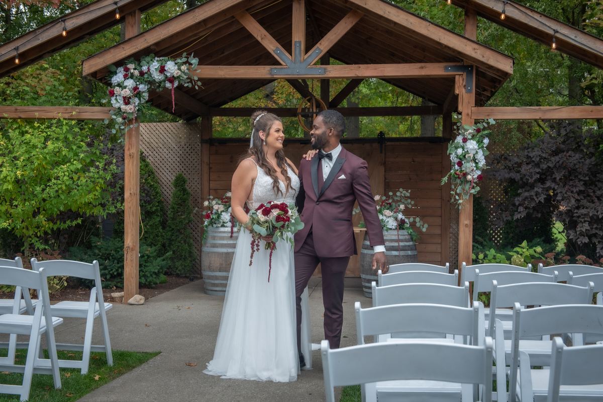 Alexandria and Derado stand beneath a rustic wooden arbor adorned with floral arrangements, smiling at each other joyfully.