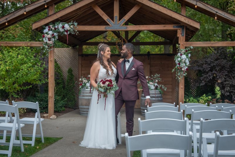 Alexandria and Derado stand beneath a rustic wooden arbor adorned with floral arrangements, smiling at each other joyfully.