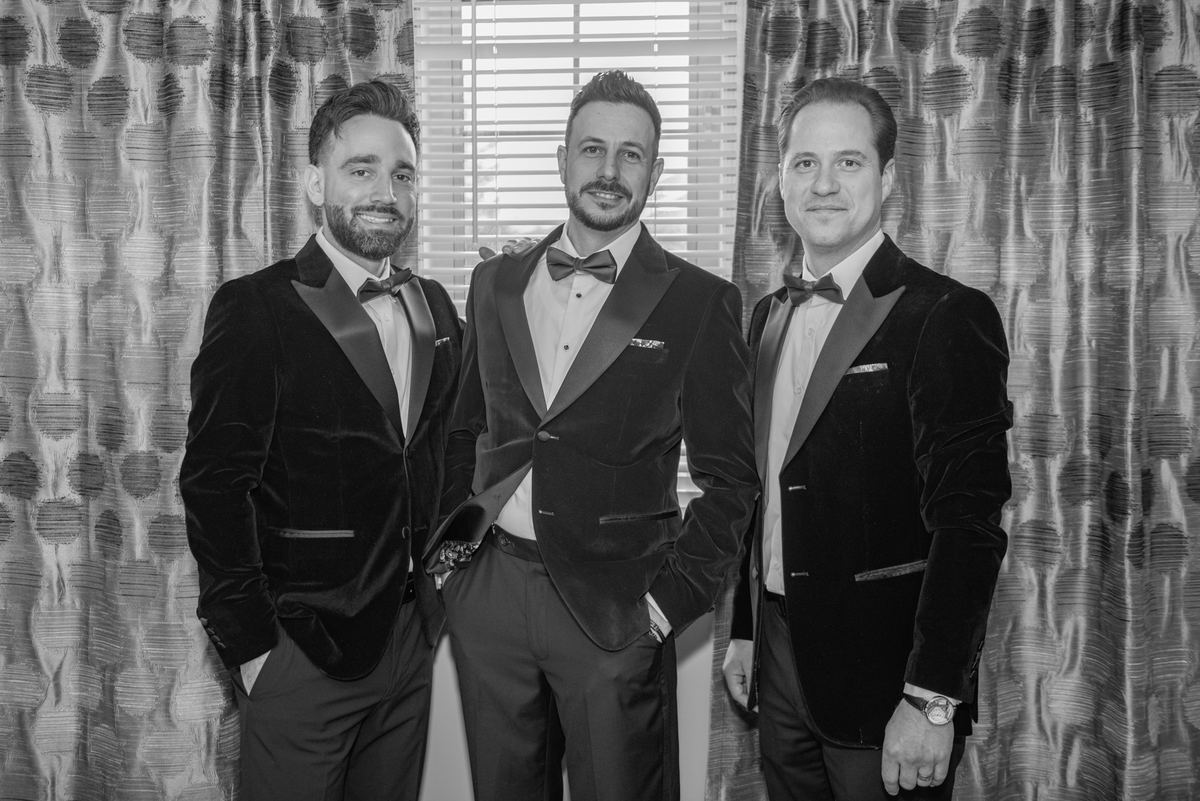 Three men in matching velvet tuxedos with bow ties stand smiling indoors, framed by patterned curtains.