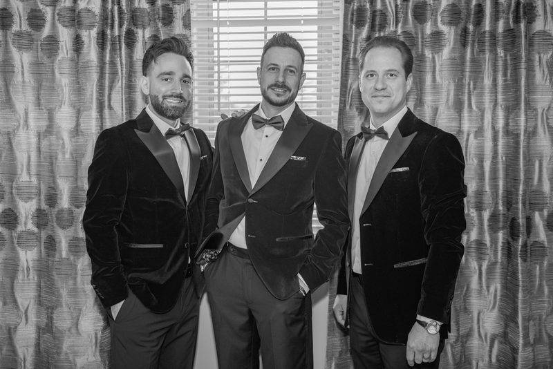 Three men in matching velvet tuxedos with bow ties stand smiling indoors, framed by patterned curtains.