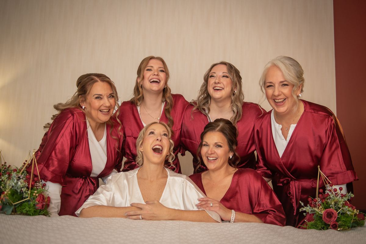 Six bridesmaids in satin burgundy robes laugh together, with the bride seated at center, under soft warm lighting.