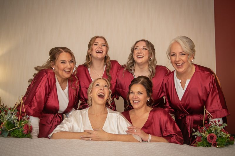 Six bridesmaids in satin burgundy robes laugh together, with the bride seated at center, under soft warm lighting.