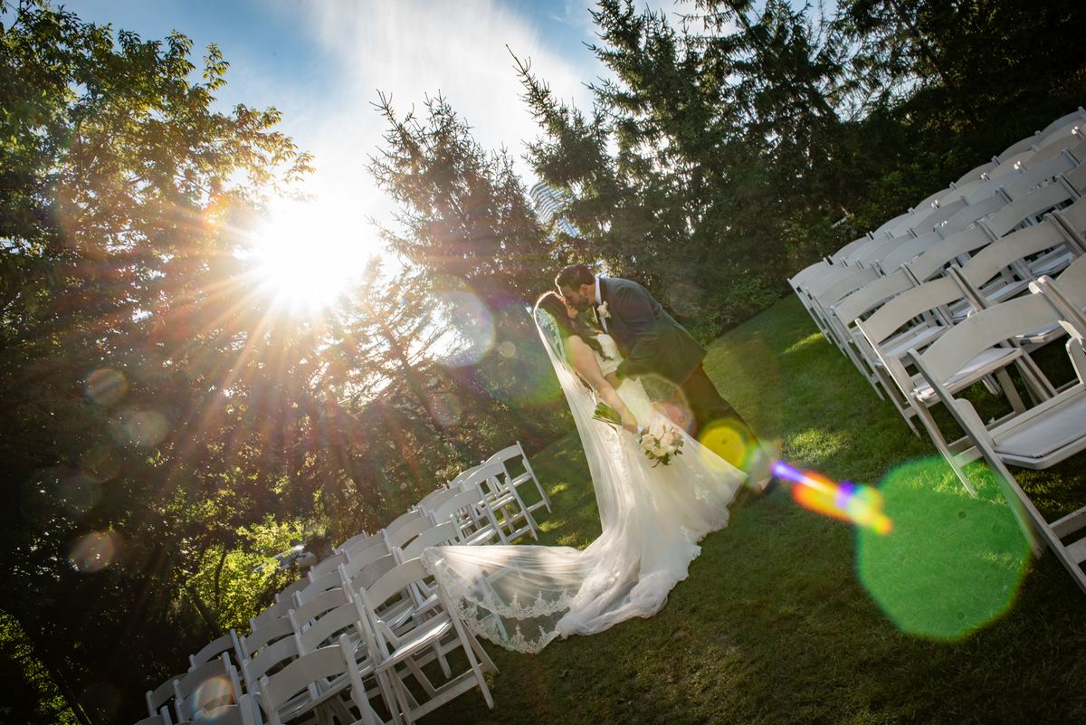 Bride and groom kiss in front of white chairs under golden sunlight filtering through trees, with lens flare.