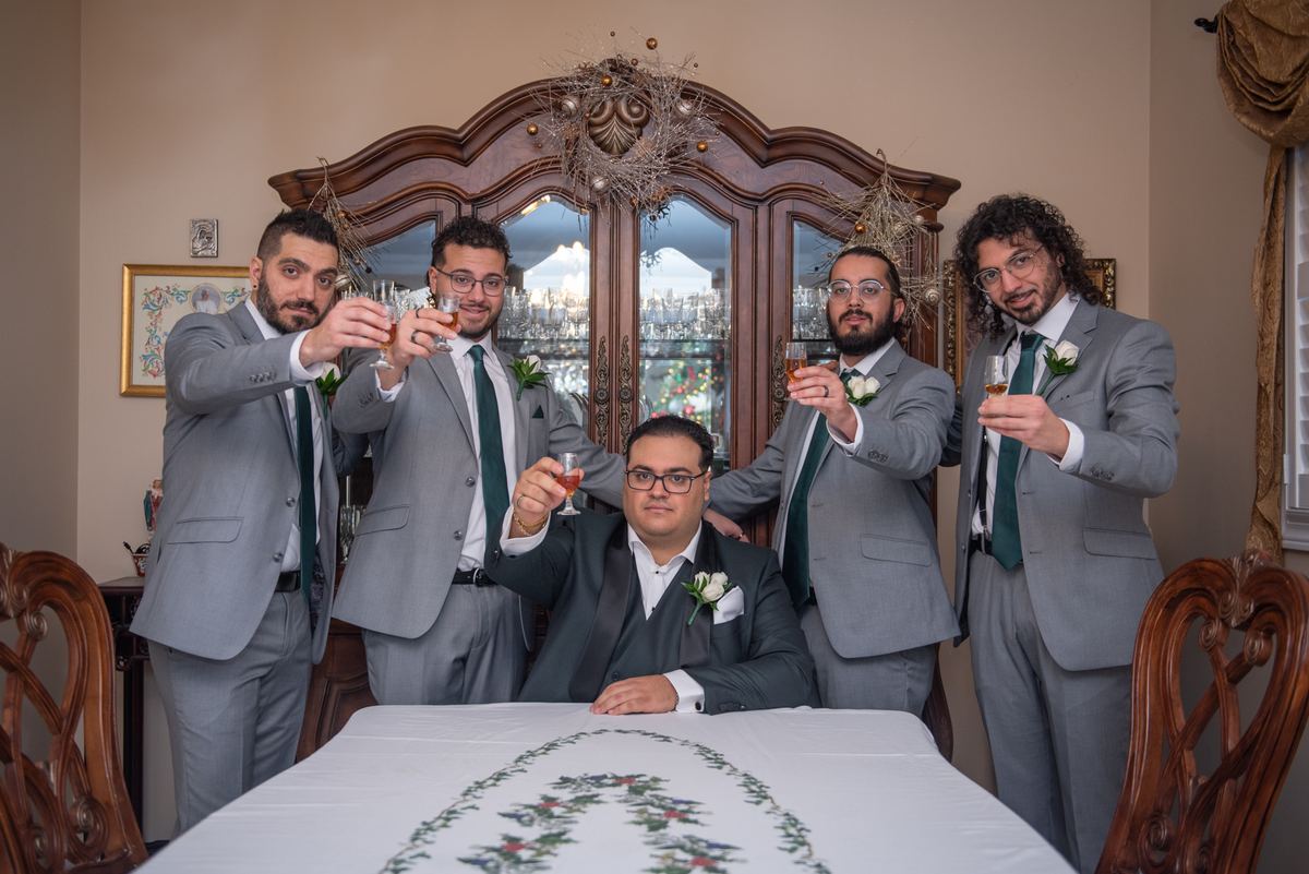 Five men, including a seated groom in dark suit and four standing groomsmen in light gray, raise glasses indoors.