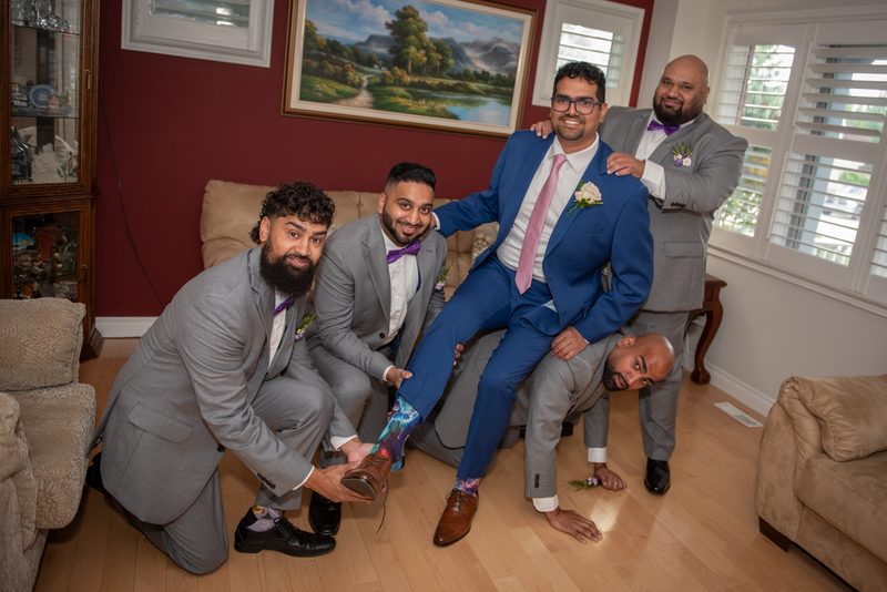 Groom in blue suit sits on couch as friends pose around him playfully, set against red wall with pastoral painting.
