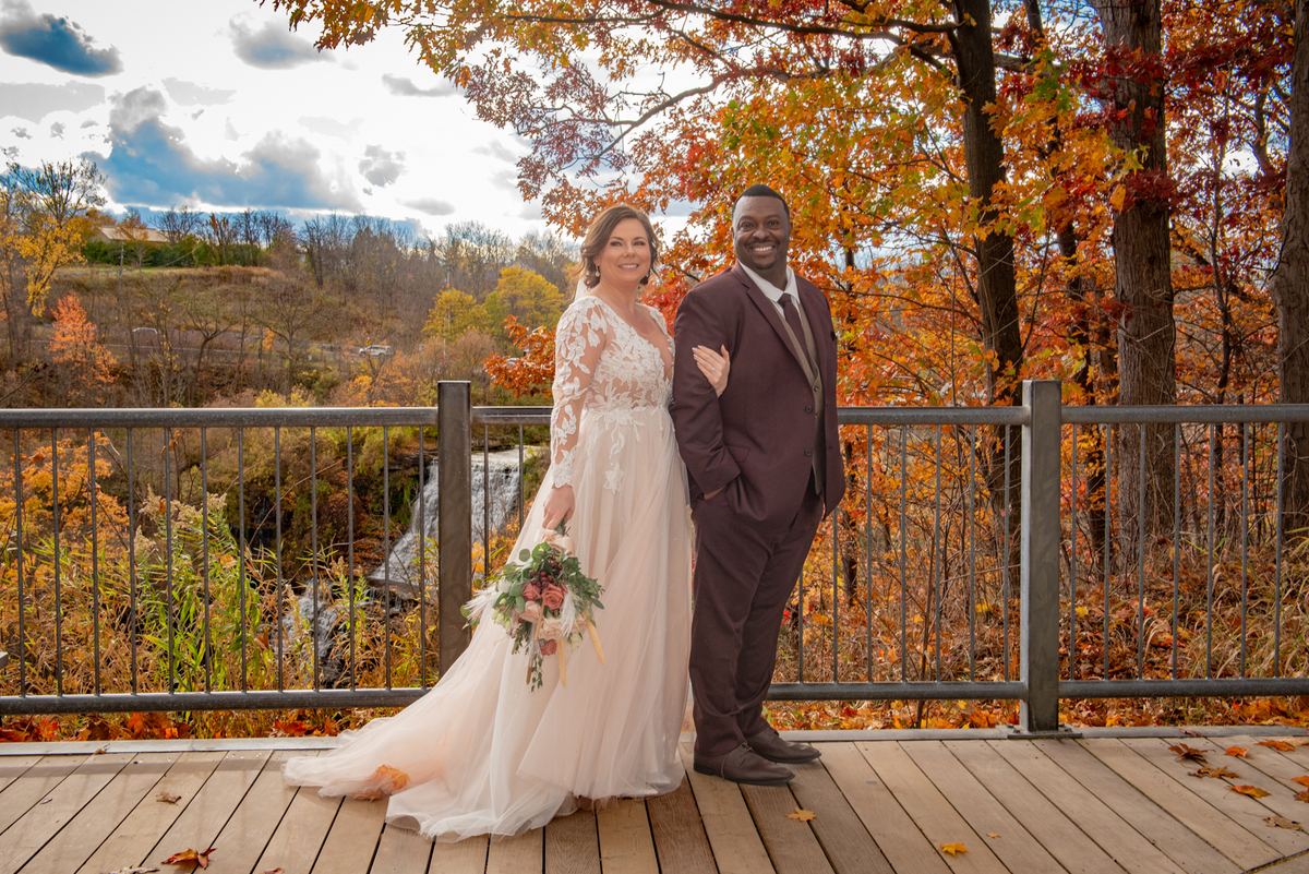 A smiling bride and groom in wedding attire stand on a wooden deck, surrounded by autumn foliage and a waterfall.