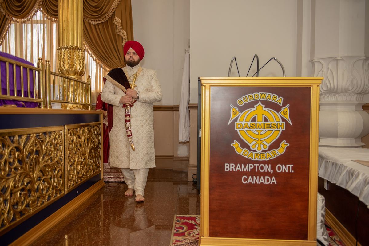 A Sikh man in ceremonial attire walks barefoot through an ornate gurdwara interior, lit by soft ambient light.