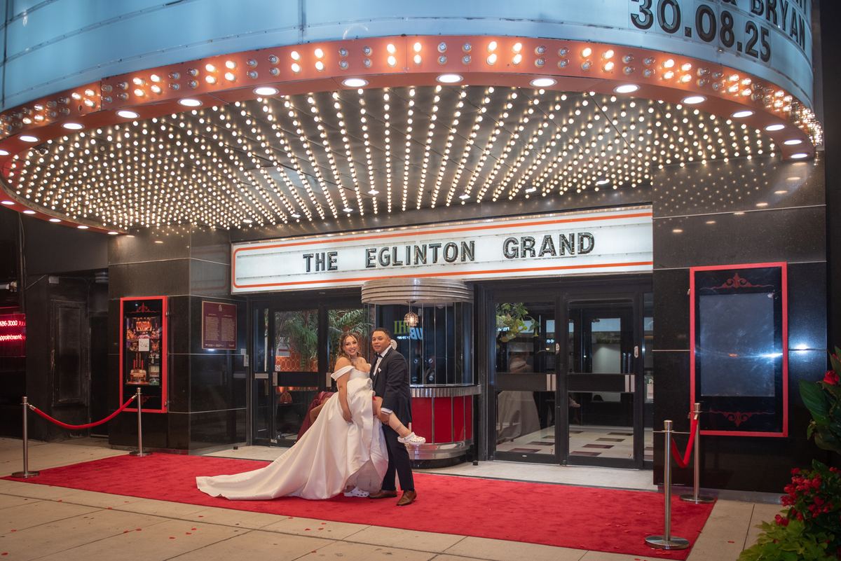 A bride and groom pose on a red carpet outside an ornate theater with glowing bulbs casting a warm light.