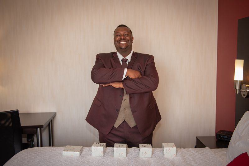 A man in a maroon suit stands with arms crossed beside a bed where five small white boxes are arranged in a row.