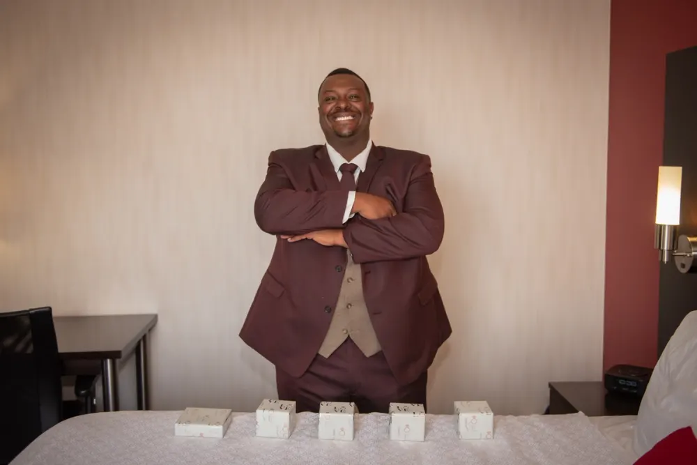 A man in a maroon suit stands with arms crossed beside a bed where five small white boxes are arranged in a row.