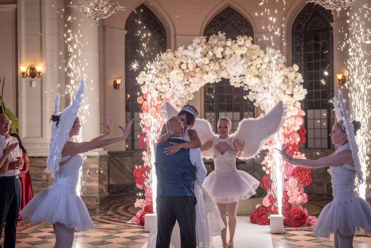 A bride and groom embrace beneath a floral arch as sparklers cascade around them in an opulent hall.