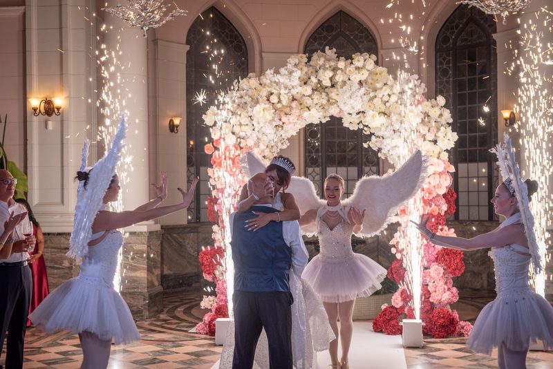 A bride and groom embrace beneath a floral arch as sparklers cascade around them in an opulent hall.