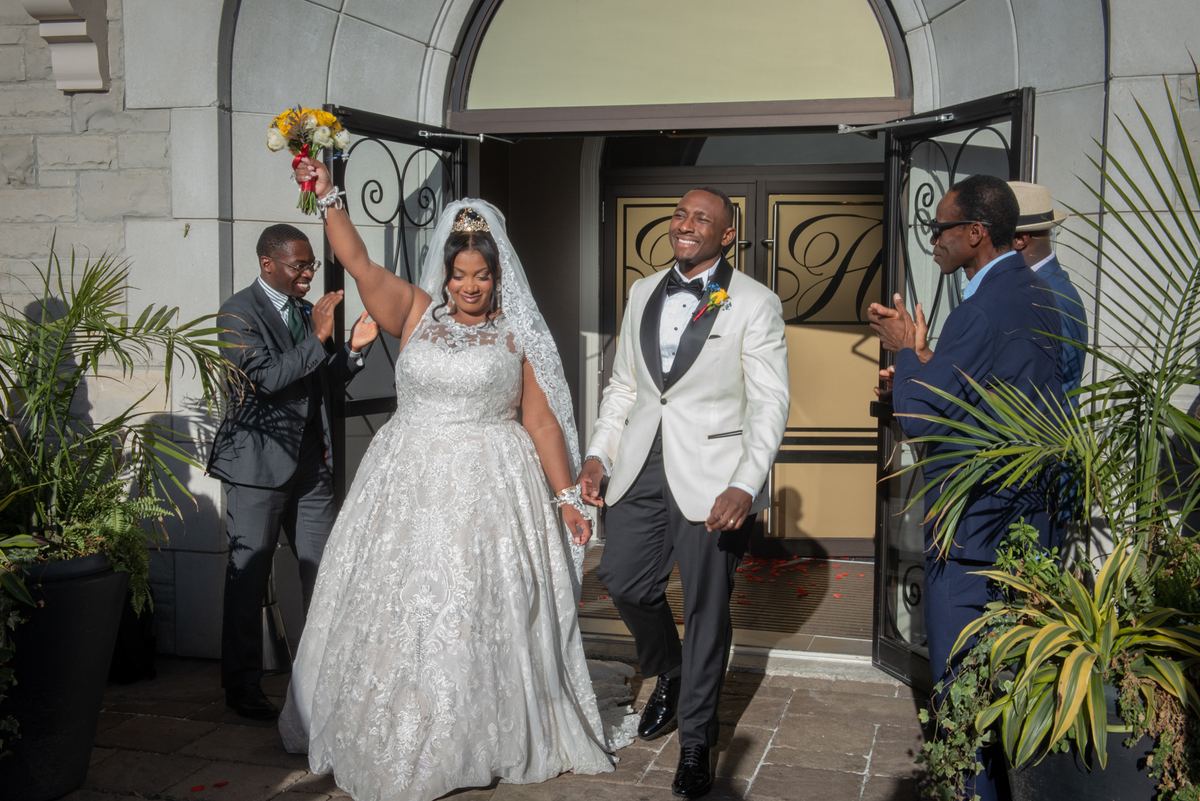 A smiling bride and groom exit a building hand-in-hand, surrounded by clapping guests in formal attire.