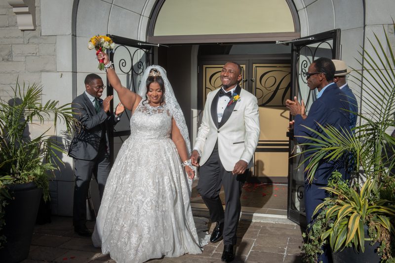 A smiling bride and groom exit a building hand-in-hand, surrounded by clapping guests in formal attire.