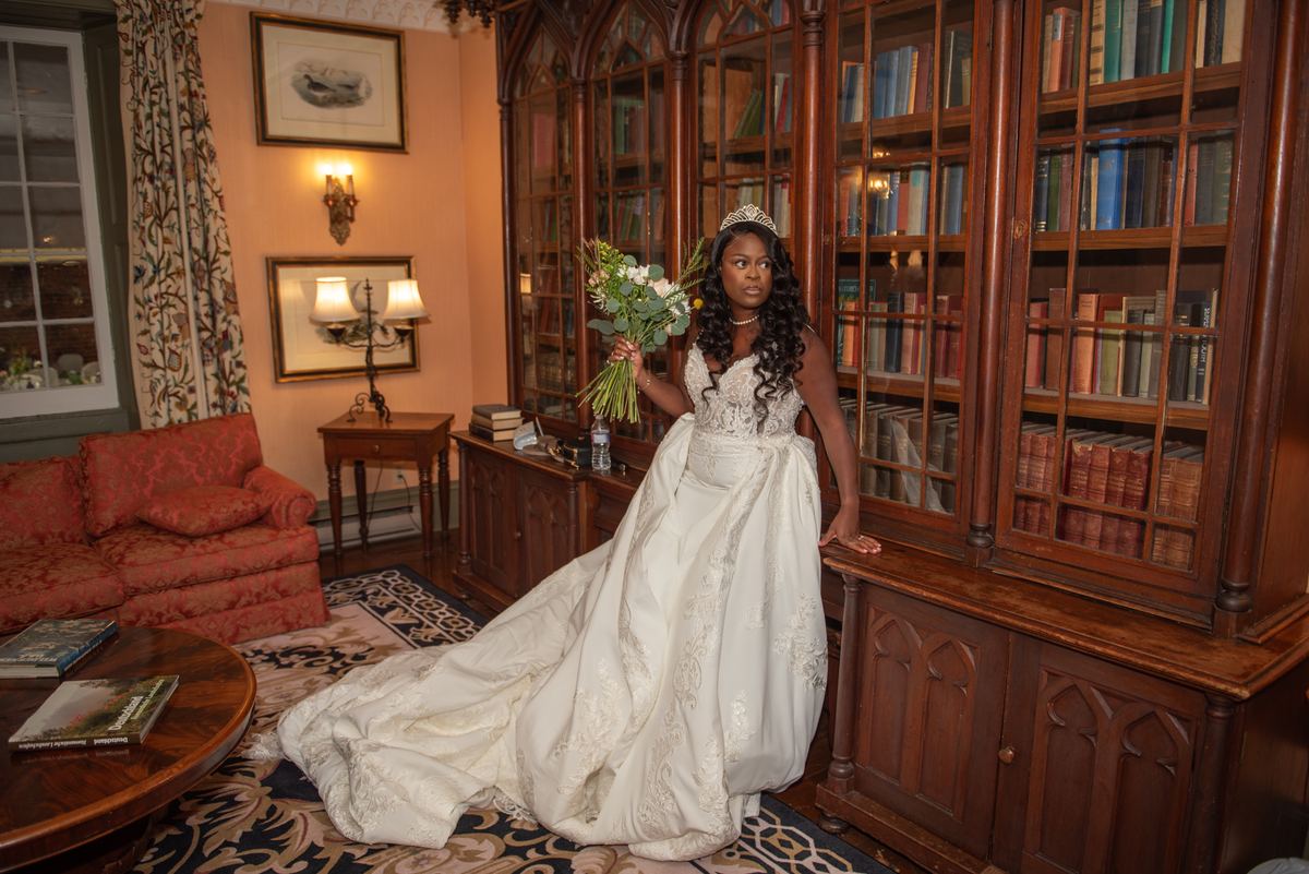 A bride in a white gown stands beside an ornate bookcase in a historic library, holding white flowers.