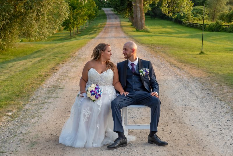 A bride in a white lace gown and groom in a navy suit sit closely on a white chair by a gravel road, surrounded by greenery.