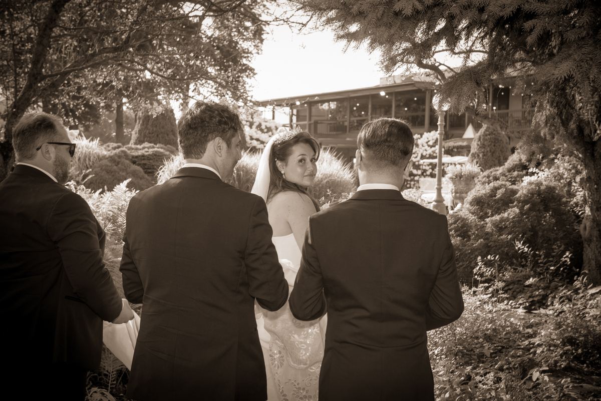 A bride in a lace gown smiles at the camera as three men in suits walk beside her in an outdoor garden setting.