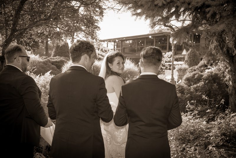 A bride in a lace gown smiles at the camera as three men in suits walk beside her in an outdoor garden setting.