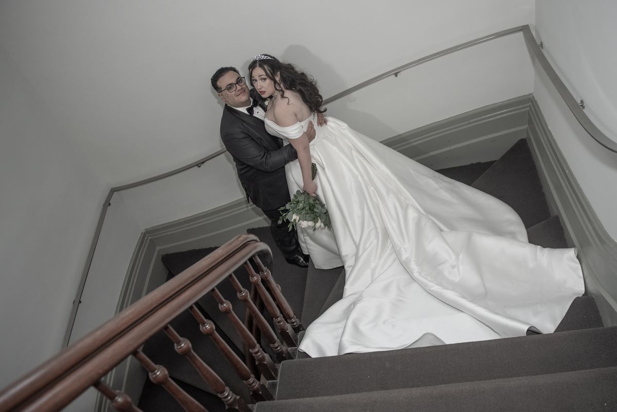 Bride and groom ascending carpeted staircase, bride holding bouquet and looking at camera, soft lighting highlights gown.