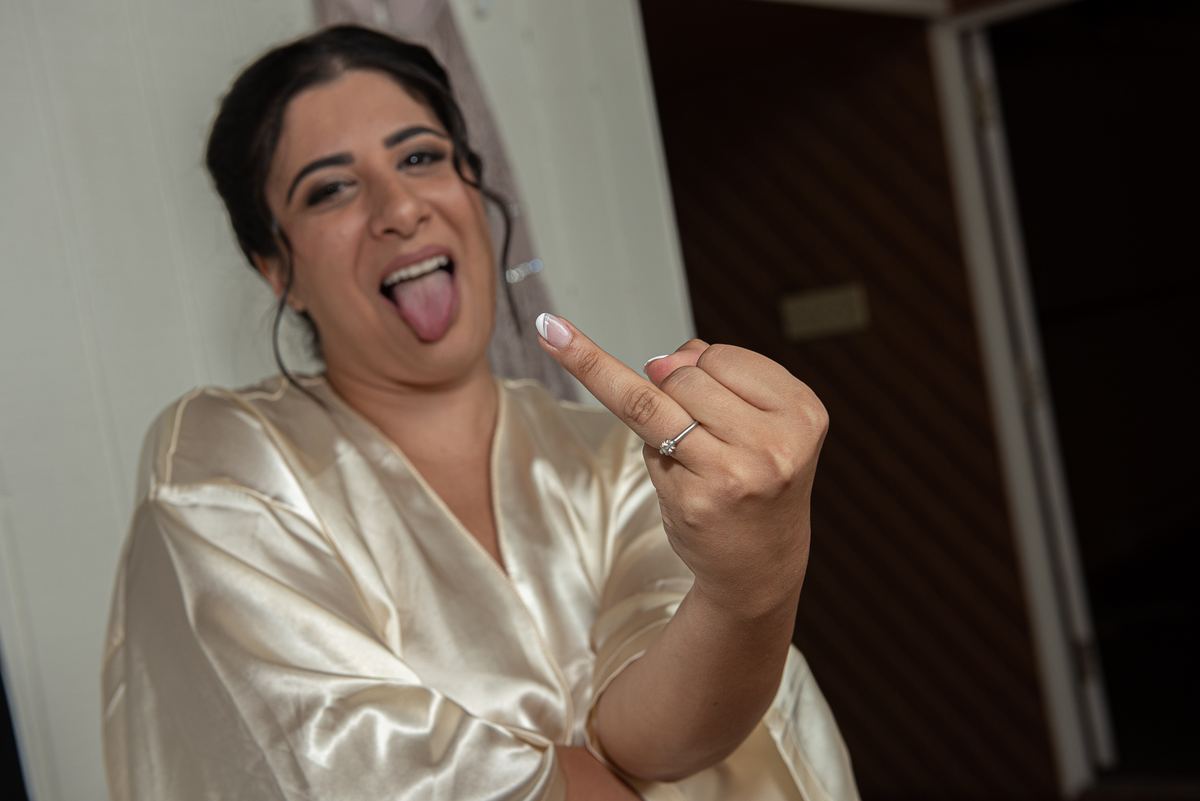 A bride in a satin robe sticks out her tongue, showing off her engagement ring against a wood-paneled background.