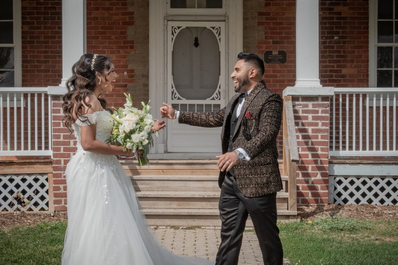 Fazana and Aman stand on the steps of a brick house, smiling and playfully reaching toward each other in their formal attire.