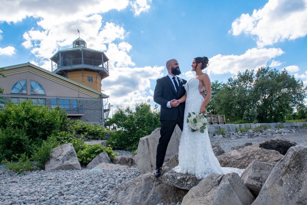 A smiling bride and groom stand on rocks by a gravel shoreline, gazing at each other near a towered building.