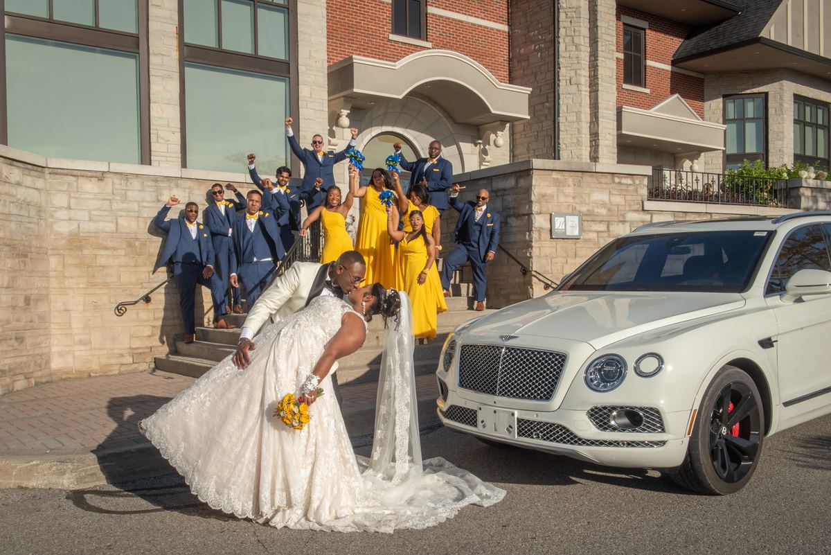 A bride and groom kiss playfully in front of a white Bentley, surrounded by cheering guests on stone steps.
