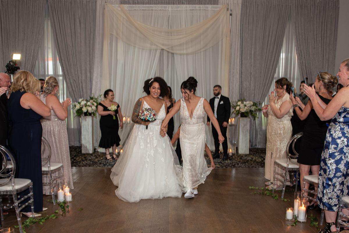 Two brides walk down the aisle hand-in-hand, smiling as guests applaud and candles along the wooden floor cast a warm glow.