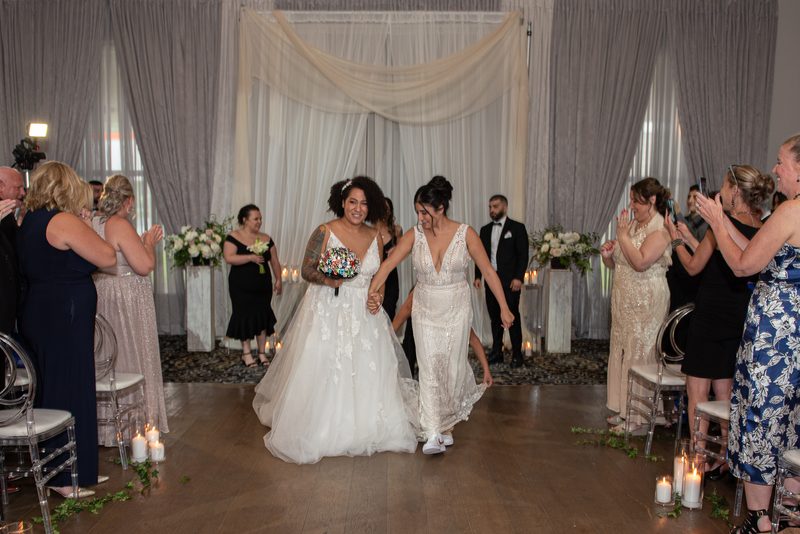 Two brides walk down the aisle hand-in-hand, smiling as guests applaud and candles along the wooden floor cast a warm glow.