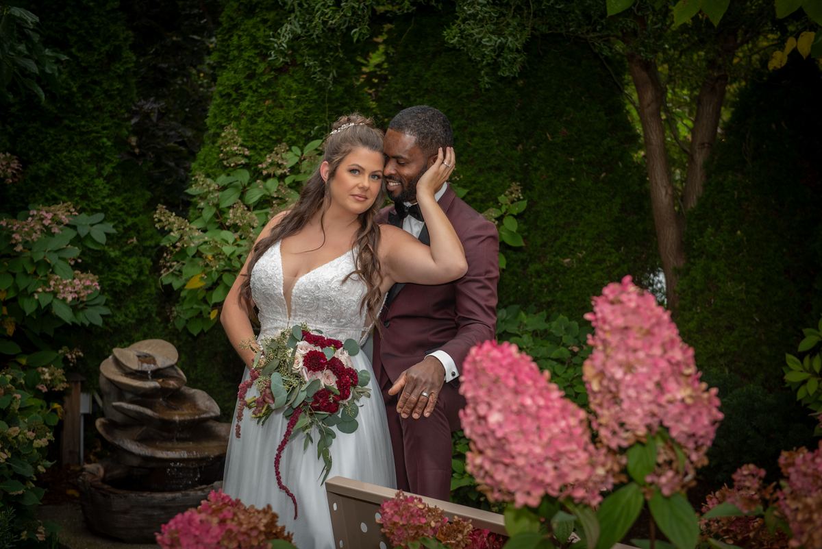 A bride in a white gown and floral bouquet stands close to her groom in a garden, hand resting on his cheek.